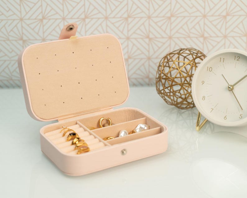 May include: A pink faux leather jewelry box with a hinged lid and multiple compartments for storing rings, earrings, and other small jewelry items. The box has a gold-colored clasp and is open to reveal the compartments.