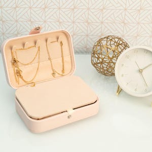 May include: A pink faux leather jewelry box with a gold chain necklace and earrings inside. The box is open and the interior is lined with a soft pink fabric. A gold wire sphere and a white clock with gold hands are in the background.