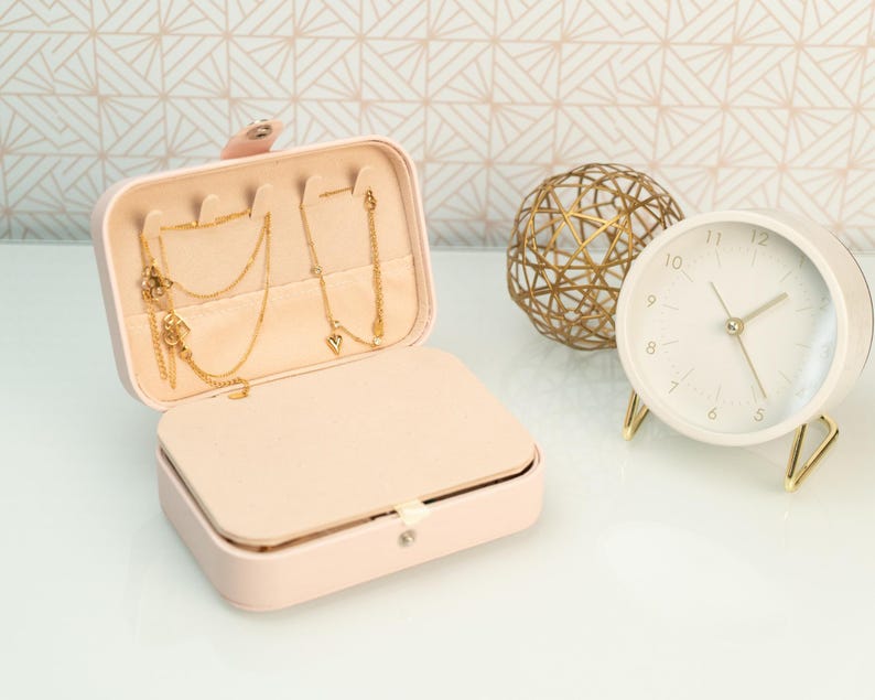 May include: A pink faux leather jewelry box with a gold chain necklace and earrings inside. The box is open and the interior is lined with a soft pink fabric. A gold wire sphere and a white clock with gold hands are in the background.