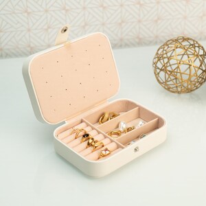 May include: A white jewelry box with a beige interior and multiple compartments for storing rings, earrings, and other jewelry. The box is open and the compartments are filled with gold jewelry.