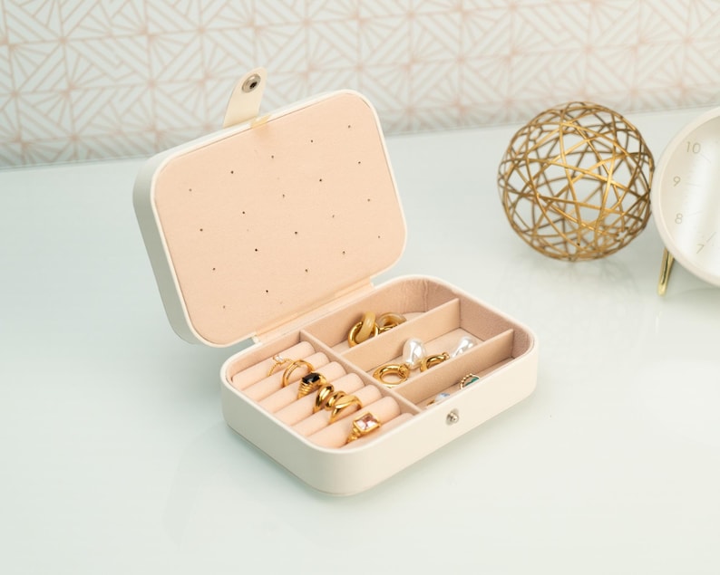 May include: A white jewelry box with a beige interior and multiple compartments for storing rings, earrings, and other jewelry. The box is open and the compartments are filled with gold jewelry.