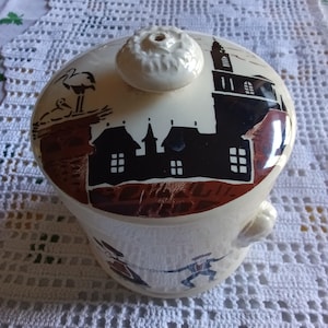 Può includere: Un barattolo in ceramica color crema con coperchio abbinato dotato di pomello. Il coperchio mostra la sagoma nera di un villaggio con una chiesa e una cicogna nel nido. La base presenta un'illustrazione colorata di due figure che danzano.