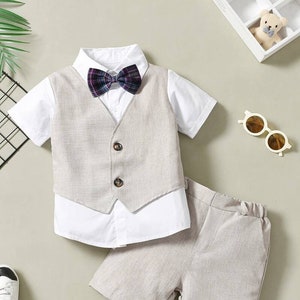 Könnte beinhalten: Ein dreiteiliges Baby-Outfit für Jungen in hellbeige. Das Outfit besteht aus einem kurzärmeligen Hemd mit Knöpfen und einer karierten Fliege, einer passenden Weste und einer Hose.