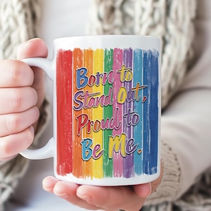 May include: White ceramic mug with a rainbow-colored vertical stripe design. The mug features the text "Born to Stand Out, Proud to Be Me" in a colorful, stylized font. The mug is held in a person's hands.