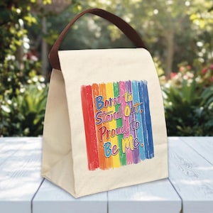 May include: A beige lunch bag with a brown handle. The bag features a rainbow-colored stripe design and the text "Born to Stand Out, Proud to Be Me" in a colorful font. The bag is sitting on a white table.