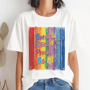 May include: White t-shirt with a rainbow-coloured vertical stripe design. The shirt features the text "Born to Stand Out, Proud to Be Me" in a colourful, stylised font. The shirt is made of a soft material.