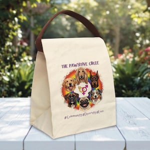 May include: A beige canvas lunch bag with a brown handle. The bag features a colorful graphic of various dog breeds inside a rainbow circle, with the text "THE PAWSITIVE CIRCLE" and hashtags #Community #Diversity #Love.