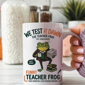 May include: White ceramic mug with a humorous design featuring a cartoon frog in glasses holding books and a coffee cup. The mug has text that reads "WE TEST AT DAWN THE TEACHER FROG" and "FUNNY TEACHER FROG".