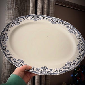 May include: An oval, off-white serving platter with a scalloped edge. The rim features a blue and black floral design. The platter is being held by a person wearing a green sweatshirt.