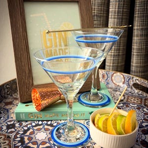 May include: Two clear martini glasses with blue rims and bases sit on a patterned tray, alongside a small bowl of citrus slices. A framed print with the text "GIN MADE ME DO IT" and a copper cocktail jigger are also present.
