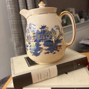 May include: A cream-coloured ceramic teapot featuring a blue and gold Asian landscape design. The teapot has a lid and gold trim. It rests on two books with dark covers.