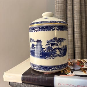 May include: A white ceramic jar with a lid, featuring a blue and white Willow pattern design. The jar has a cylindrical shape with a gold trim. The design includes scenes of bridges, pagodas, and landscapes. The jar is sitting on top of a stack of books.