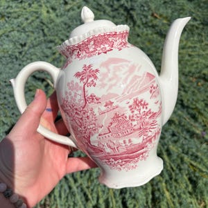May include: A white ceramic teapot with a red landscape design. The teapot has a curved spout, a handle, and a lid with a decorative finial. The design features buildings, trees, and figures, creating a scenic view.