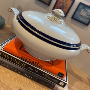 Peut inclure: Une soupière en céramique blanche avec couvercle, ornée d'une bande bleu marine et de détails dorés. La soupière repose sur une pile de livres, dont "Little Book of Hermes" et "The Dress". Poignées décoratives et un bouton.