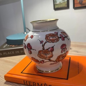 Puede incluir: Un jarrón de porcelana blanca con un borde dorado y un diseño floral. El jarrón presenta un delicado patrón de flores rosas y naranjas con tallos y hojas verdes.