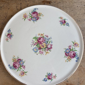 May include: A round, off-white serving tray with a gold rim. The tray is decorated with clusters of pink, purple, blue, and yellow flowers. The floral pattern is arranged in a symmetrical design across the surface of the tray.