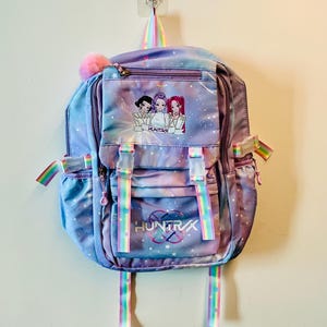 May include: A pastel purple and blue galaxy-print backpack featuring a front pocket with a graphic of three anime-style characters and the text HUNTRX. The bag includes rainbow-striped webbing straps, white plastic buckles, and a pink pom-pom.