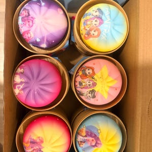 May include: Six round, pleated containers arranged in a cardboard box. Each lid features a colorful gradient design with printed cartoon character illustrations and the text "HUNTRIX" on some, showcasing various shades of pink, purple, blue, and yellow.