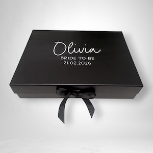 May include: A black gift box tied with a black ribbon. The lid is inscribed with the name "Olivia" in a cursive font, and the text "BRIDE TO BE" and the date "21.02.2026" in a simple font.