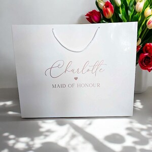 May include: White gift bag with a white rope handle. The bag has the name "Charlotte" written in rose gold script with a small heart and the text "Maid of Honour" in rose gold.