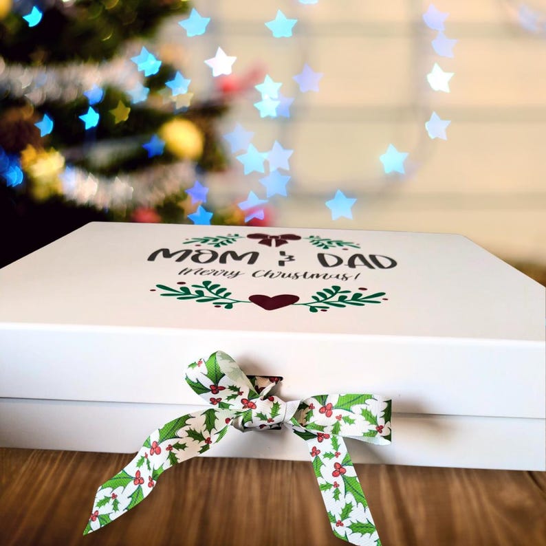 May include: A white gift box with the text "Mom & Dad Merry Christmas!" in black and green. The box is tied with a white ribbon decorated with green holly leaves and red berries. The background features a blurred Christmas tree with star-shaped lights.
