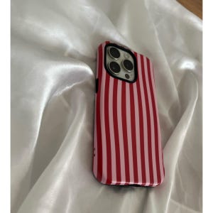 Op de afbeelding: Een rood en roze gestreept telefoonhoesje. De hoes heeft een glanzende afwerking en een zwarte rand. De cameralenzen zijn zichtbaar aan de bovenkant. De hoes ligt op een witte, zijdeachtige ondergrond.