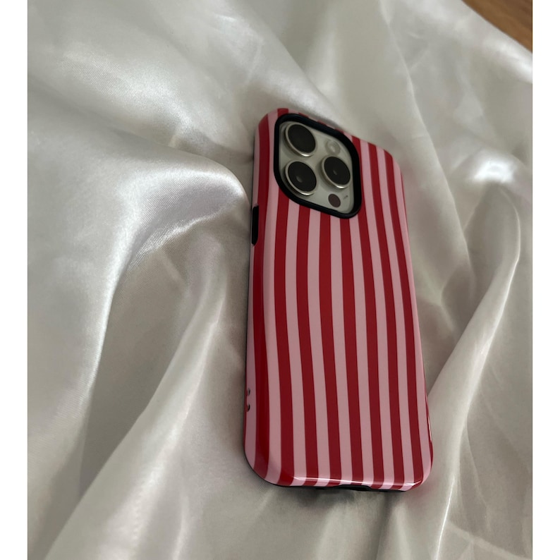 Op de afbeelding: Een rood en roze gestreept telefoonhoesje. De hoes heeft een glanzende afwerking en een zwarte rand. De cameralenzen zijn zichtbaar aan de bovenkant. De hoes ligt op een witte, zijdeachtige ondergrond.