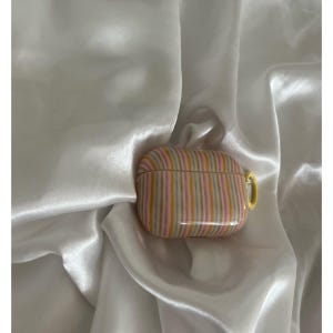 May include: A striped AirPods Pro case in shades of pink, yellow, and gray. The case has a yellow loop for attaching to a keychain. The case is resting on a white, silky fabric.