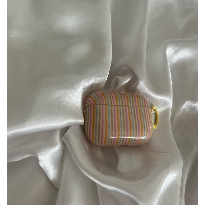 May include: A striped AirPods Pro case in shades of pink, yellow, and gray. The case has a yellow loop for attaching to a keychain. The case is resting on a white, silky fabric.