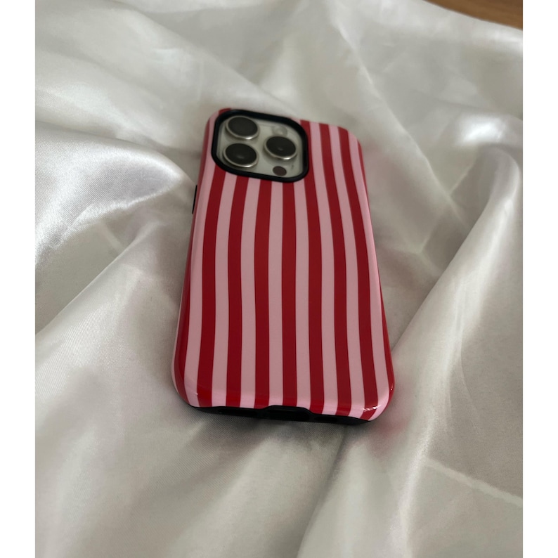 Op de afbeelding: Een rood-roze gestreepte telefoonhoes. De hoes heeft een glanzende afwerking en een zwarte rand. De telefoonhoes ligt op een witte, zijdeachtige ondergrond.