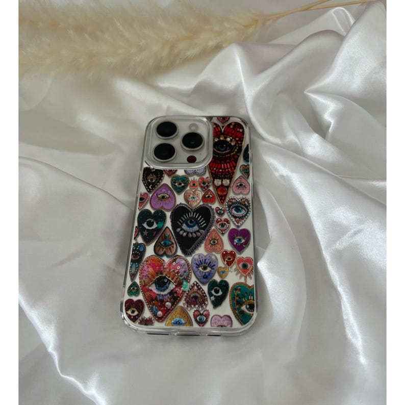 May include: Clear phone case with a colorful heart design. The hearts feature eye motifs and various patterns in shades of red, purple, blue, and green. The case has a clear border and covers the phone's back.