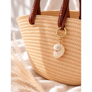 Könnte beinhalten: Eine geflochtene Strohtasche mit dunkelbraunen Ledergriffen und einem goldfarbenen Metallringverschluss. Ein weißer spiralförmiger Muschelanhänger hängt an dem Ring und setzt einen natürlichen Akzent auf dem strukturierten, hellbeigen Flechtmaterial.