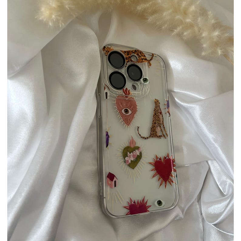 May include: A clear phone case with a colorful design featuring a heart with an eye, a leopard, and other decorative elements.