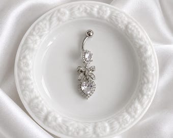 May include: A silver-toned navel ring featuring a curved barbell with a round clear stone at the top, a central bow-shaped charm encrusted with small clear stones, and a dangling heart-shaped pendant set with a large clear stone and a halo of smaller stones.