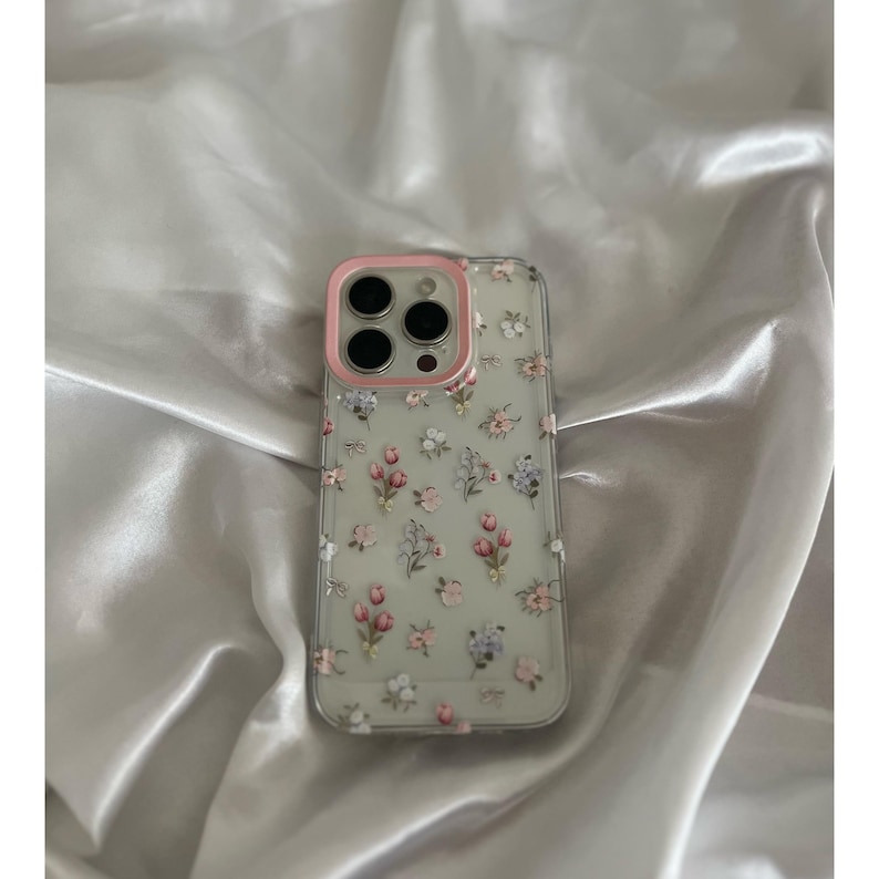 Pode incluir: Capa de telefone transparente com um padr&atilde;o floral com flores rosa, roxas e brancas. A capa tem uma borda rosa ao redor das lentes da c&acirc;mera.