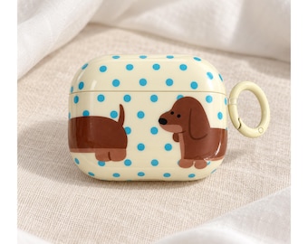 May include: A cream-colored protective case for wireless earbuds featuring a repeating pattern of small blue polka dots and a brown dachshund illustration. The case includes a hinged lid and a light-colored circular carabiner attached to the side.