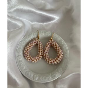May include: A pair of teardrop-shaped earrings. The earrings are made of small, light pink and cream-colored beads. They are arranged in a teardrop shape and have gold-colored hooks. The earrings are displayed on a white, decorative plate.