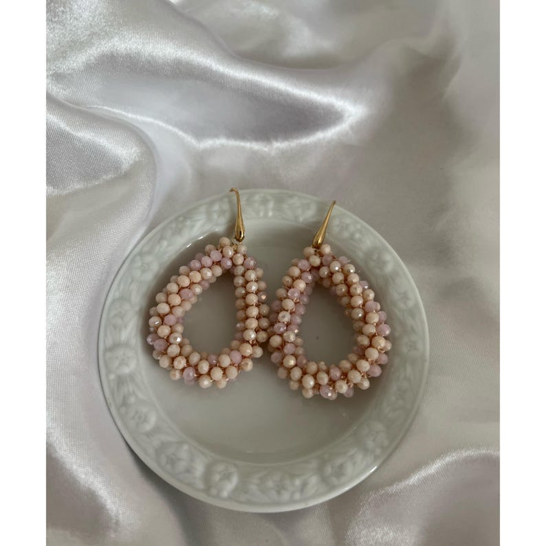 May include: A pair of teardrop-shaped earrings. The earrings are made of small, light pink and cream-colored beads. They are arranged in a teardrop shape and have gold-colored hooks. The earrings are displayed on a white, decorative plate.