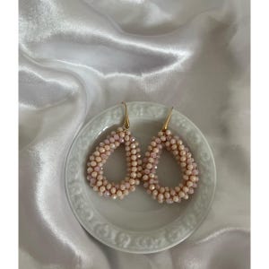 May include: A pair of teardrop-shaped earrings with a beaded design. The earrings are light pink and beige, with gold-colored hooks. They are displayed on a white, decorative plate.