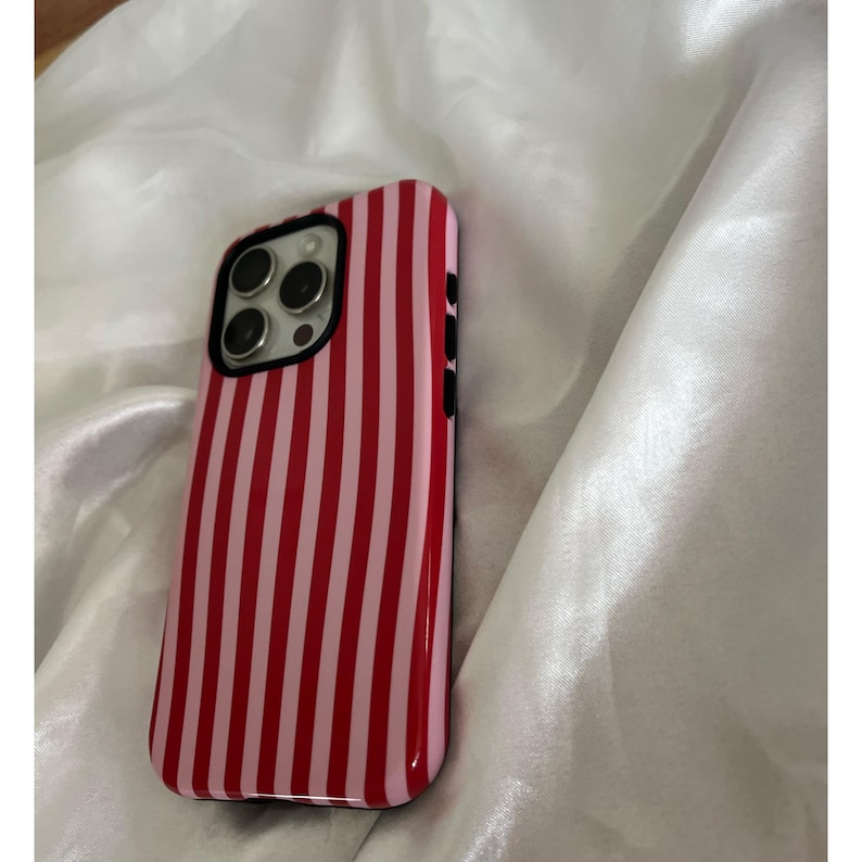 Op de afbeelding: Een rood-roze gestreepte telefoonhoes. De hoes heeft een glanzende afwerking en een zwarte rand rond de camera. De hoes is ontworpen voor een smartphone en heeft uitsparingen voor de camera en knoppen.