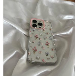 Pode incluir: Capa de telefone transparente com uma moldura rosa para a c&acirc;mera, adornada com um delicado padr&atilde;o floral de flores rosa, brancas e lavanda, e pequenos la&ccedil;os. A capa est&aacute; sobre um tecido de cetim branco.
