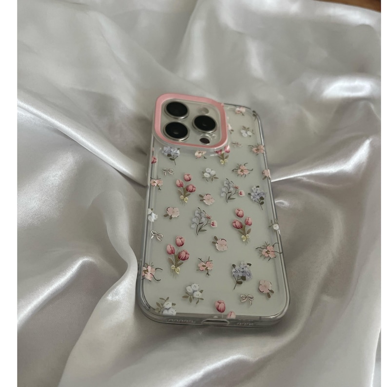 Pode incluir: Capa de telefone transparente com uma moldura rosa para a c&acirc;mera, adornada com um delicado padr&atilde;o floral de flores rosa, brancas e lavanda, e pequenos la&ccedil;os. A capa est&aacute; sobre um tecido de cetim branco.