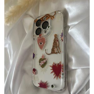 May include: A clear phone case with a white background and colorful designs. The designs include a heart with an eye, a leopard, and a heart with flowers. The case is for an iPhone with a triple lens camera.