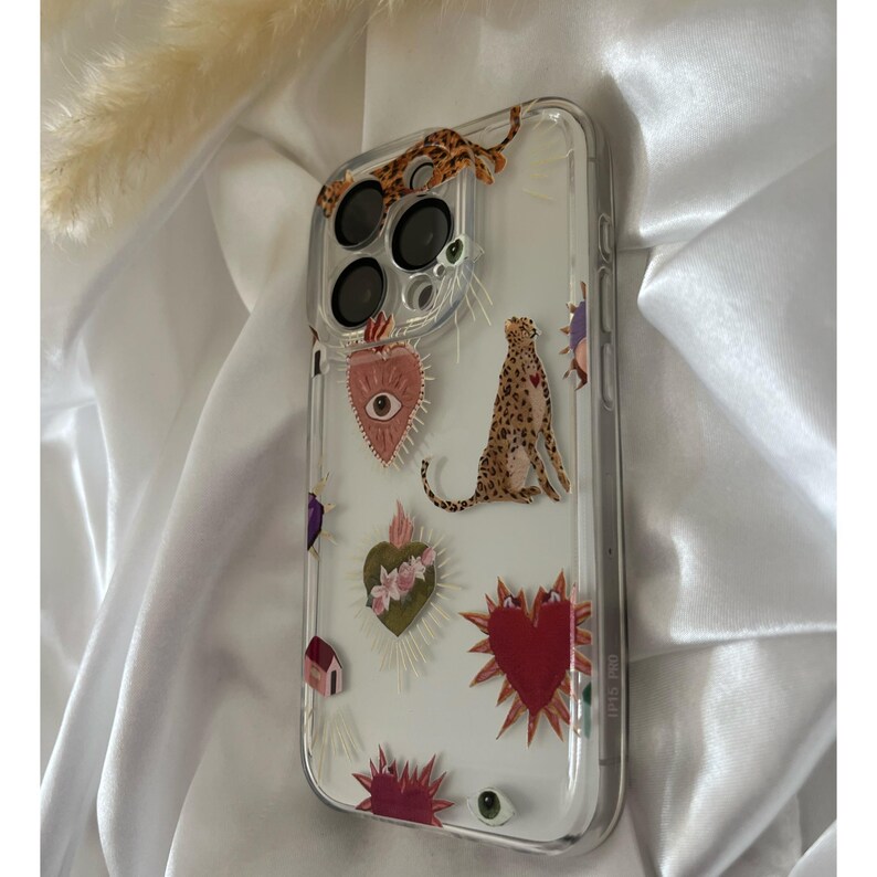May include: A clear phone case with a white background and colorful designs. The designs include a heart with an eye, a leopard, and a heart with flowers. The case is for an iPhone with a triple lens camera.