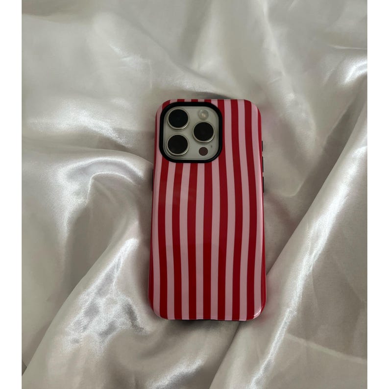 Op de afbeelding: Een rood en roze gestreept telefoonhoesje met een zwarte rand. De hoes is ontworpen voor een smartphone en heeft een uitsparing voor de camera. De hoes ligt op een witte satijnen ondergrond.