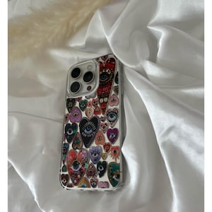 May include: A clear phone case featuring a colorful heart and eye pattern. The design includes various heart shapes in red, purple, and teal, each with an eye motif. The case is set against a white satin background.