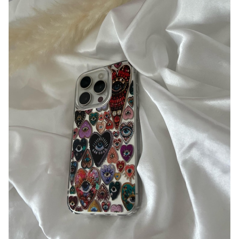 May include: A clear phone case featuring a colorful heart and eye pattern. The design includes various heart shapes in red, purple, and teal, each with an eye motif. The case is set against a white satin background.
