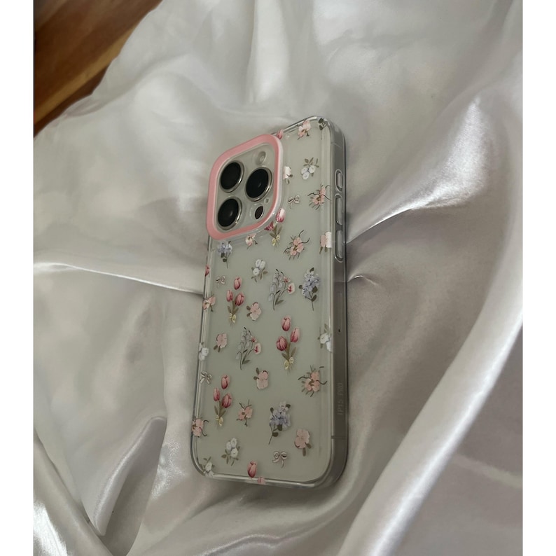 Pode incluir: Capa de telefone transparente com um padr&atilde;o floral com flores rosa, roxas e brancas. A capa tem um anel de c&acirc;mera rosa e foi projetada para um smartphone. A capa est&aacute; em uma superf&iacute;cie de cetim branco.
