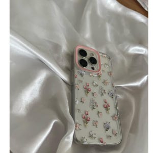 Pode incluir: Capa de telefone transparente com um anel de c&acirc;mera rosa e um padr&atilde;o floral de flores rosa, roxas e brancas. A capa est&aacute; sobre um tecido de cetim branco.