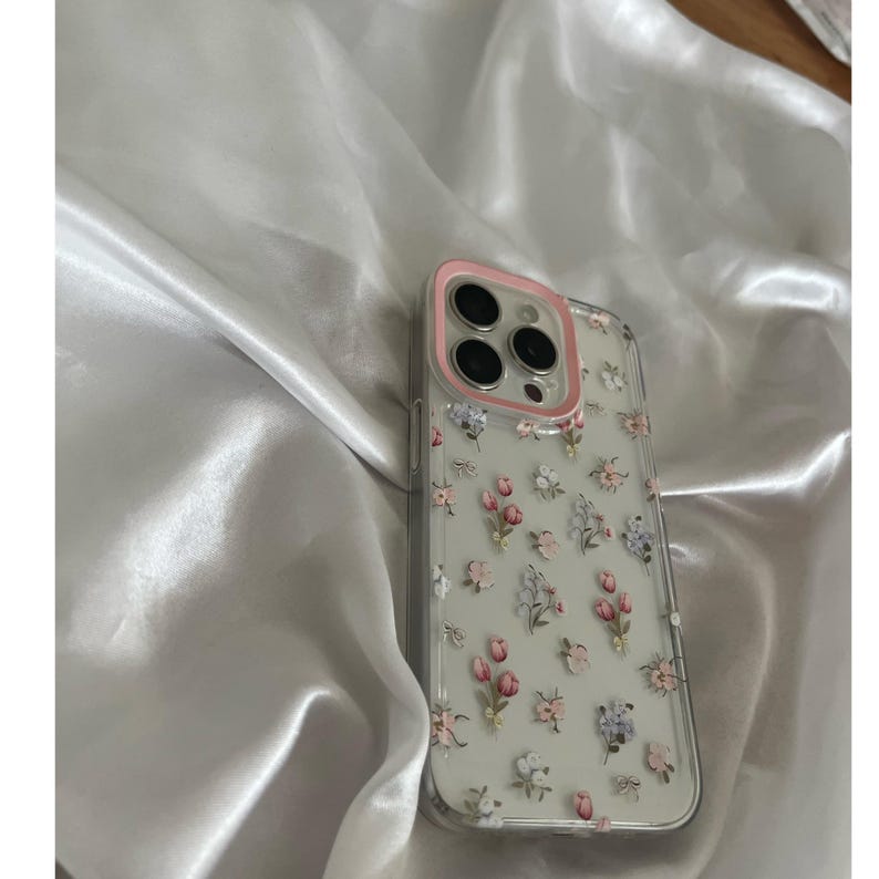 Pode incluir: Capa de telefone transparente com um anel de c&acirc;mera rosa e um padr&atilde;o floral de flores rosa, roxas e brancas. A capa est&aacute; sobre um tecido de cetim branco.